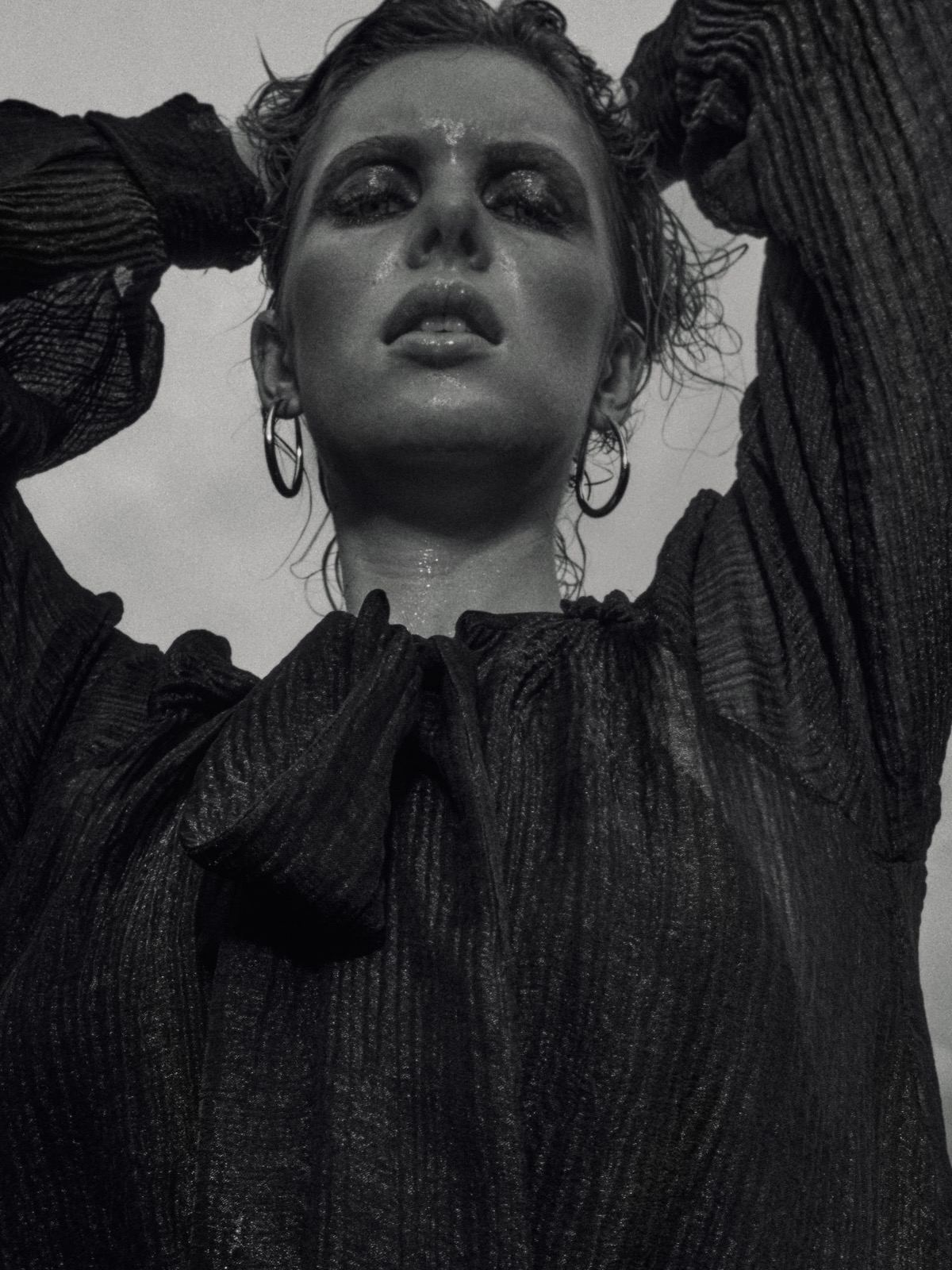 Model Caitlin Sanderson in black and white, wet-look styling, arms raised in a dark textured blouse, photographed by Eli Samuel for Latest Magazine. Wardrobe by Matt Taylor, make-up by Leigh Dempsey-McCoy, hair by Michelle Eileen, with Caitlin Sanderson represented by Chic Management.
