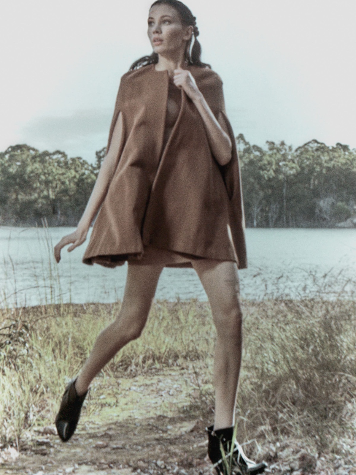Model Jess Tanner mid-stride beside a lake in a tan cape and black boots, July 2017, photographed by Eli Samuel. Wardrobe by Seoyoung We, make-up by Lucy Whelan, hair by Chevelle Sullivan-Pont, with Jess Tanner represented by Que Models.